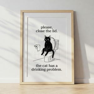 May include: A framed print featuring a black and white illustration of a black cat sitting on a toilet. The text reads "please, close the lid. the cat has a drinking problem."