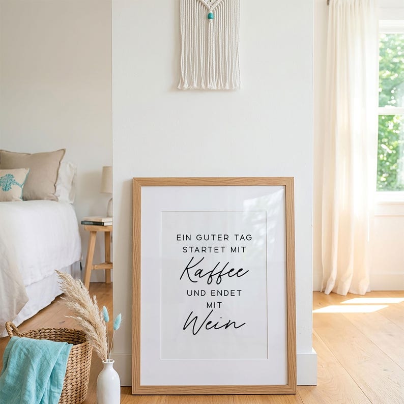 May include: A framed print with the text "EIN GUTER TAG STARTET MIT Kaffee UND ENDET MIT Wein" in black script. The frame is light brown, and the print is displayed in a bright, airy room with a bed and a window.