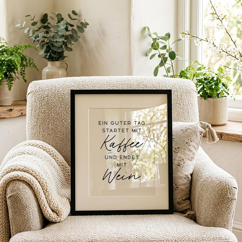 May include: A framed print with a black border and a cream-colored mat. The print features the German text "Ein guter Tag startet mit Kaffee und endet mit Wein" in a stylish font. The print is displayed on a beige armchair.