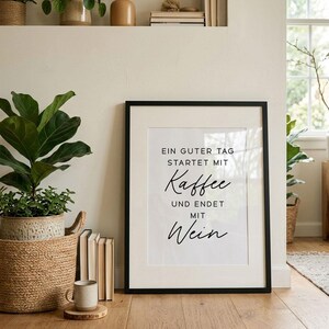 May include: A framed print with the text "EIN GUTER TAG STARTET MIT Kaffee UND ENDET MIT Wein" in black script. The print is in a black frame and is displayed on a wooden floor. A coffee mug sits on a wooden coaster in front of the print.