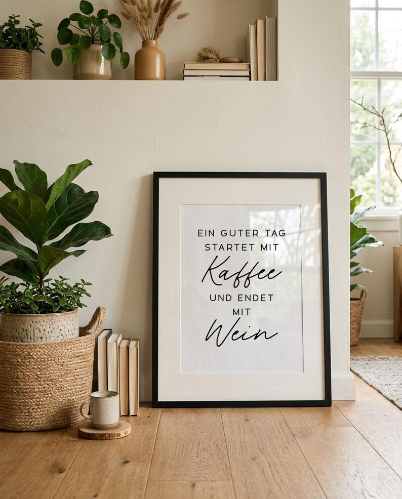 May include: A framed print with the text "EIN GUTER TAG STARTET MIT Kaffee UND ENDET MIT Wein" in black script. The print is in a black frame and is displayed on a wooden floor. A coffee mug sits on a wooden coaster in front of the print.