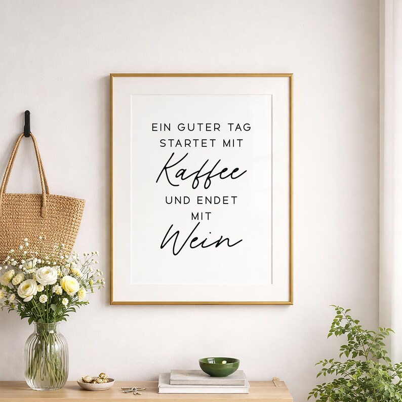May include: Framed wall art with the text "EIN GUTER TAG STARTET MIT Kaffee UND ENDET MIT Wein" in black script on a white background. The gold frame is displayed on a white wall, with a straw tote bag and flowers to the left.