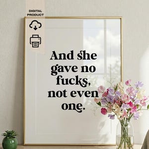 Può includere: Stampa incorniciata con la scritta "And she gave no fucks, not even one". La stampa è in una cornice dorata e si trova accanto a un vaso di fiori. Le parole sono in grassetto, in nero.
