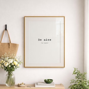 Può includere: Una stampa artistica incorniciata con il testo nero Be nice (or leave) in un carattere in stile macchina da scrivere su sfondo bianco. La stampa è racchiusa in una sottile cornice dorata e appesa a una parete sopra una superficie in legno.