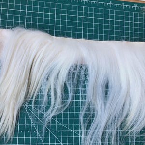 Puede incluir: Una tira larga de piel sintética blanca con fibras finas y rectas. La piel está colocada horizontalmente sobre una alfombrilla de corte verde que presenta una cuadrícula blanca con marcas numéricas del 19 al 41 en el borde izquierdo.