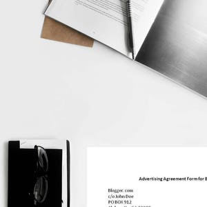 May include: A white sheet of paper with a black and white image on the right side. The left side of the paper has text about the "immeasurable shades of gray" and a pen is lying on top of the paper. The paper is on top of a brown envelope. Below the paper is a black and white notebook with a pair of black glasses on top of it. On the right side of the image is a white sheet of paper with the text "Advertising Agreement Form for Bloggers.com" and a form to fill out with the following information: Business/Person Name, Business Address, Phone Number, Email, and Authorized Person (if other than listed above).