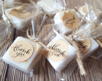 May include: Square, white soaps wrapped in clear cellophane and tied with twine. Each soap has a round, brown sticker with the words "thank you" in black script. The soaps are arranged on a light brown wooden surface.
