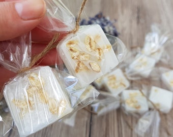 May include: Square, white soaps with visible oats are wrapped in clear cellophane and tied with twine. The soaps are arranged on a wooden surface, with some in focus and others blurred in the background. The soaps are a natural, handmade product.