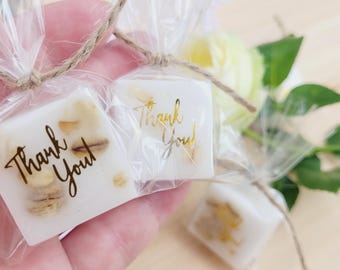 May include: Small, square, white favors with dried floral inclusions, wrapped in clear cellophane and tied with twine. Each favor has the words "Thank You" printed on it in gold or brown script. A few white and yellow roses are in the background.