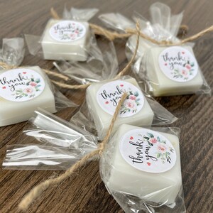 May include: Small white square soaps wrapped in clear cellophane bags with a brown twine bow. Each soap has a round sticker with the text "Thank you" and a floral design.