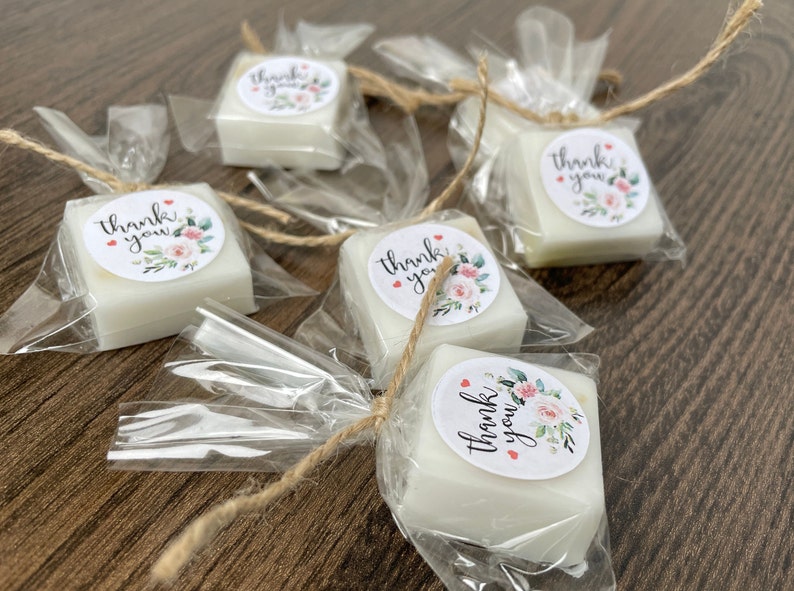 May include: Small white square soaps wrapped in clear cellophane bags with a brown twine bow. Each soap has a round sticker with the text "Thank you" and a floral design.