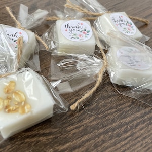 May include: A collection of small, white, square-shaped soaps wrapped in clear cellophane and tied with twine. Some soaps have decorative elements like gold flakes or oatmeal. The soaps have small, round, white labels with the text "Thank you" printed on them.