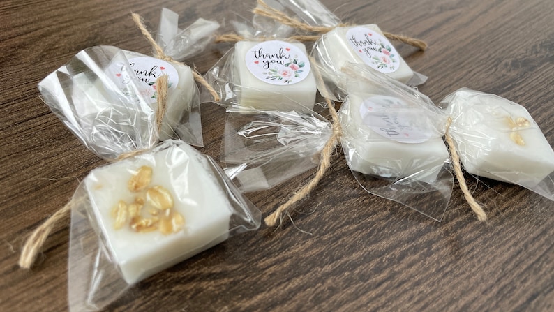 May include: A collection of small, white, square-shaped soaps wrapped in clear cellophane and tied with twine. Some soaps have decorative elements like gold flakes or oatmeal. The soaps have small, round, white labels with the text "Thank you" printed on them.