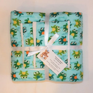 May include: A light blue flannel baby blanket and two matching burp cloths featuring a repeating pattern of green frogs. The set is tied with a white ribbon and includes a tag that reads: Flannel Baby Blanket with 2 burp cloths, Size: 30" x 30", 100% cotton flannel with polyester batting, Machine wash in warm water, Dry on low heat.