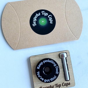 Op de afbeelding: Een houten doos met een zwarte fiets-deksel en een zilveren bout. Het deksel heeft de tekst "Keep pedalling Dad! You've got this!" en de doos is gelabeld met "Bespoke Top Caps".
