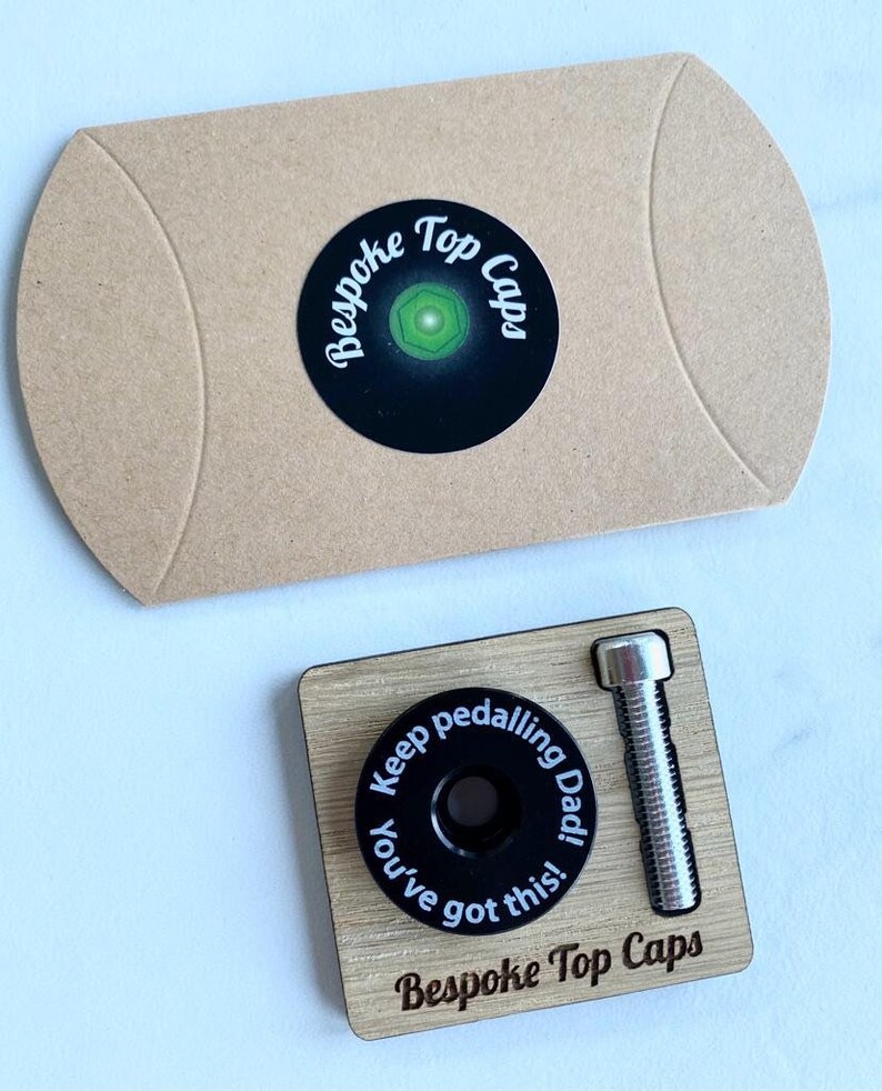 Op de afbeelding: Een houten doos met een zwarte fiets-deksel en een zilveren bout. Het deksel heeft de tekst "Keep pedalling Dad! You've got this!" en de doos is gelabeld met "Bespoke Top Caps".