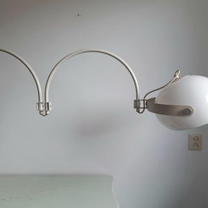 Può includere: Una lampada da parete con un lungo braccio metallico curvo color bianco sporco che forma due archi. La testa della lampada è un globo sferico in vetro bianco tenuto in posizione da una fascia metallica orizzontale, collegata da una cerniera.