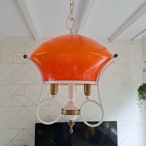 Op de afbeelding: Een witte en oranje mid-century modern hanglamp met drie lampen. De lamp heeft een ronde, oranje glazen kap en een wit metalen frame met drie ringen.