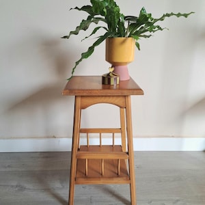 May include: A wooden side table with a rectangular top and a lower shelf. A yellow and pink planter with a green plant sits on top, along with a small decorative box. The table has a classic, vintage design.
