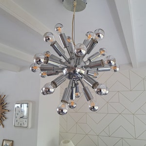 Może przedstawiać: Chromowy żyrandol szklany z wieloma żarówkami. Żyrandol ma kształt gwiazdy, a żarówki promieniują z centrum.