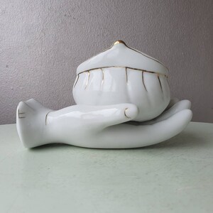 May include: White porcelain trinket box shaped like a hand holding a small, white, gold-trimmed, lidded bowl.