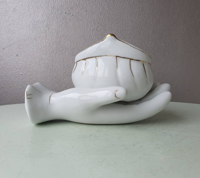 May include: White porcelain trinket box shaped like a hand holding a small, white, gold-trimmed, lidded bowl.