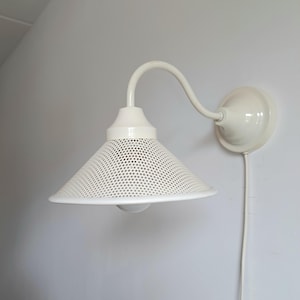Pode incluir: Luminária de parede branca com uma tela cónica perfurada. A luminária tem um braço curvo e uma base redonda. O design é simples e moderno. A lâmpada é montada numa parede branca.