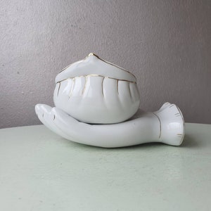 May include: White ceramic hand holding a white ceramic bowl with gold trim. The bowl has a fluted design.