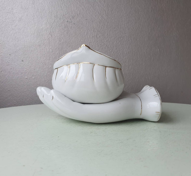 May include: White ceramic hand holding a white ceramic bowl with gold trim. The bowl has a fluted design.