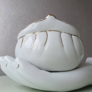 May include: White ceramic trinket dish with a gold rim and a hand-shaped base. The dish is shaped like a shell and has a smooth, glossy finish.