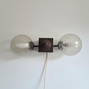 Könnte beinhalten: Eine Wandleuchte mit einem quadratischen, dunkelbraunen Holzsockel und einem zentralen Knopf. Zwei schwarze Metallarme ragen horizontal hervor und tragen jeweils einen kugelförmigen, klaren Glasschirm mit einer dezenten, gesprenkelten Textur.