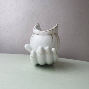 May include: White ceramic hand-shaped planter with a gold rimmed bowl on top. The planter is designed to look like a hand with four fingers.
