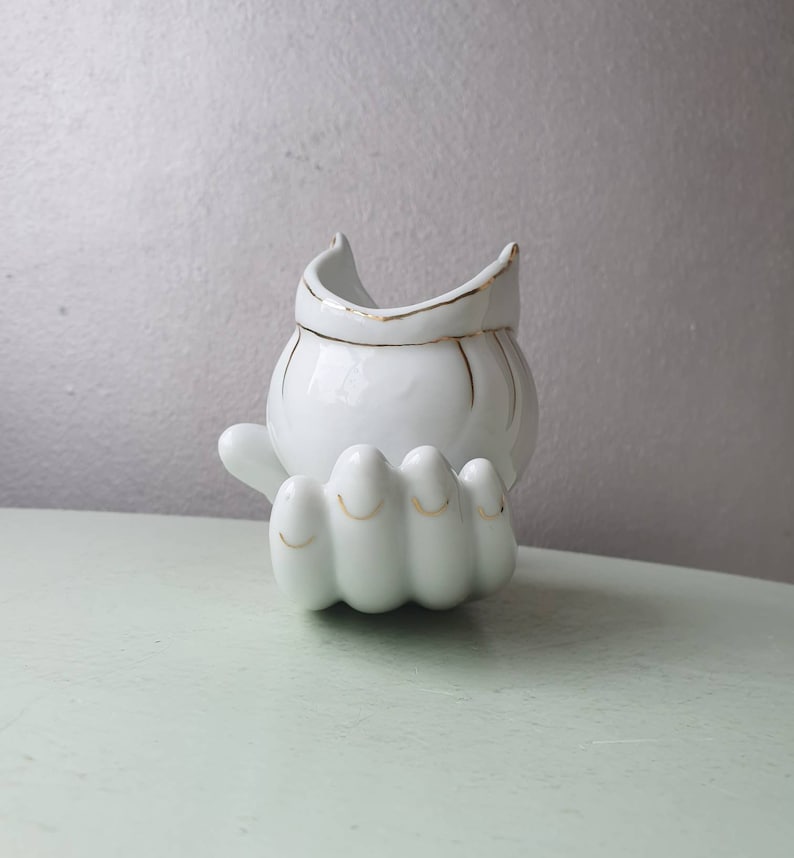May include: White ceramic hand-shaped planter with a gold rimmed bowl on top. The planter is designed to look like a hand with four fingers.