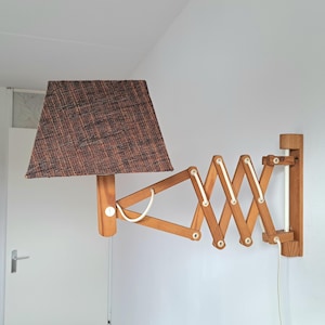 May include: A wooden wall-mounted lamp with an accordion-style arm and a brown lampshade. The lamp is made of light-colored wood and has a white cord. The lampshade is a dark brown color.