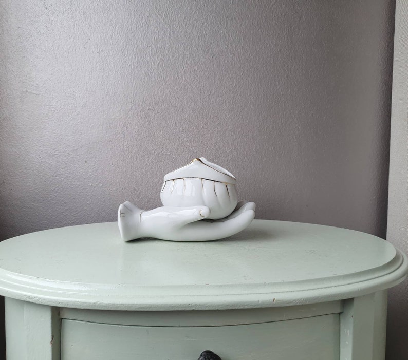 May include: White porcelain trinket box shaped like a hand holding a small, white, lidded bowl with gold trim.