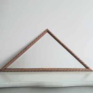 May include: A triangular mirror with a wooden frame. The frame has a textured, ridged design and a warm brown color. The mirror is resting on a white surface against a white wall.