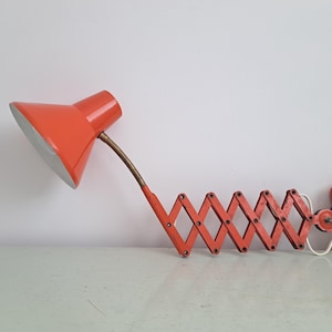 Könnte beinhalten: Eine orangefarbene Schreibtischlampe im Vintage-Stil mit einem Ziehharmonika-Arm und einem konischen Lampenschirm. Die Lampe hat einen flexiblen, messingfarbenen Hals und eine Wandhalterung. Die Lampe ist aus Metall und hat ein weißes Netzkabel.