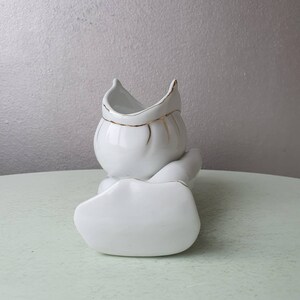 May include: White ceramic vase with a gold trim, shaped like a person sitting with legs crossed. The vase is designed to hold flowers or other decorative items.