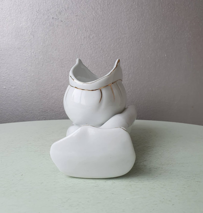 May include: White ceramic vase with a gold trim, shaped like a person sitting with legs crossed. The vase is designed to hold flowers or other decorative items.