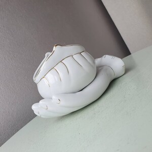 May include: White ceramic hand-shaped trinket dish with a gold trim. The dish is shaped like a hand holding a small bowl.