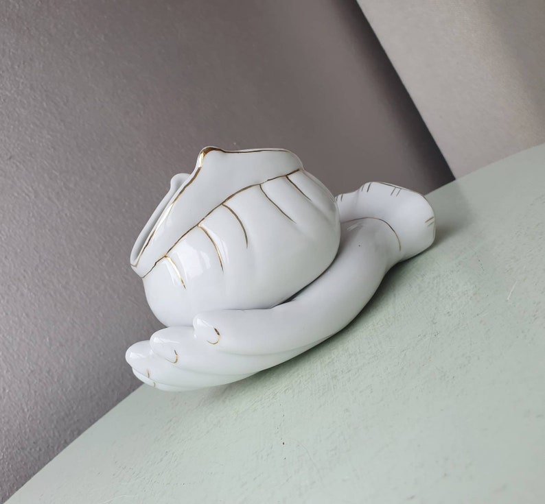 May include: White ceramic hand-shaped trinket dish with a gold trim. The dish is shaped like a hand holding a small bowl.