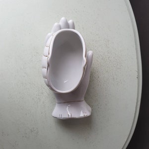 May include: White ceramic hand-shaped trinket dish with gold trim. The dish is shaped like a cupped hand with the fingers curled inwards.