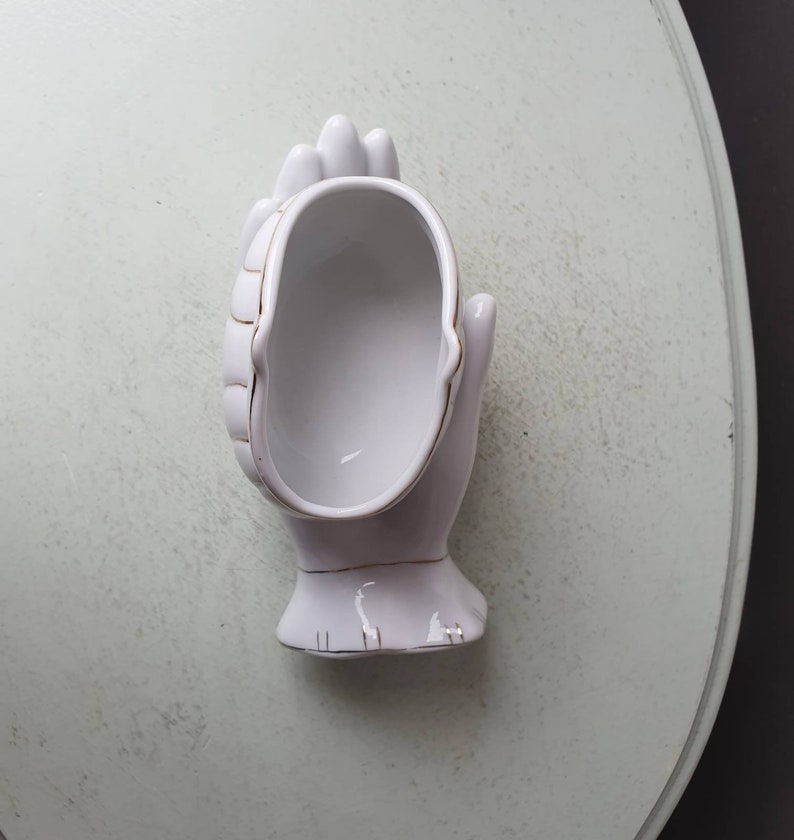 May include: White ceramic hand-shaped trinket dish with gold trim. The dish is shaped like a cupped hand with the fingers curled inwards.