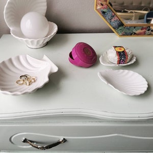 Op de afbeelding: Witte keramische schelpenvormige schalen op een witte commode met een roze compact en een stuk zeep verpakt in een gepatroneerd papiertje.