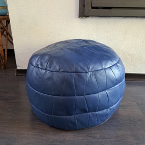 Puede incluir: Un puf redondo de cuero azul con un diseño acolchado. El puf es redondo y tiene una parte superior ligeramente aplanada.