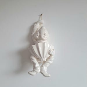 Puede incluir: Adorno de pared de cerámica blanca que representa a un payaso sonriente tocando el acordeón. El payaso lleva sombrero y botas. El acordeón tiene forma de abanico. La figura está suspendida por una cuerda.