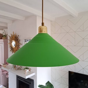Op de afbeelding: Een groene, kegelvormige hanglamp met een gouden metalen top. De lamp hangt aan een plafond in een kamer met witte muren.