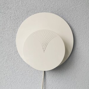 Op de afbeelding: Een witte plafondlamp met een ronde, geperforeerde diffuser. De lamp is gemonteerd op een witte muur.
