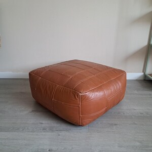 May include: A square, brown leather ottoman with a stitched, grid-like pattern. The ottoman is a warm, medium brown color and sits on a gray wooden floor. The design is simple and modern, suitable for a living room or lounge area.
