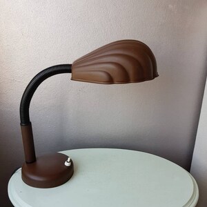 Peut inclure: Une lampe de bureau marron avec un bras noir flexible et un abat-jour en forme de coquille. La lampe a un interrupteur blanc sur la base.